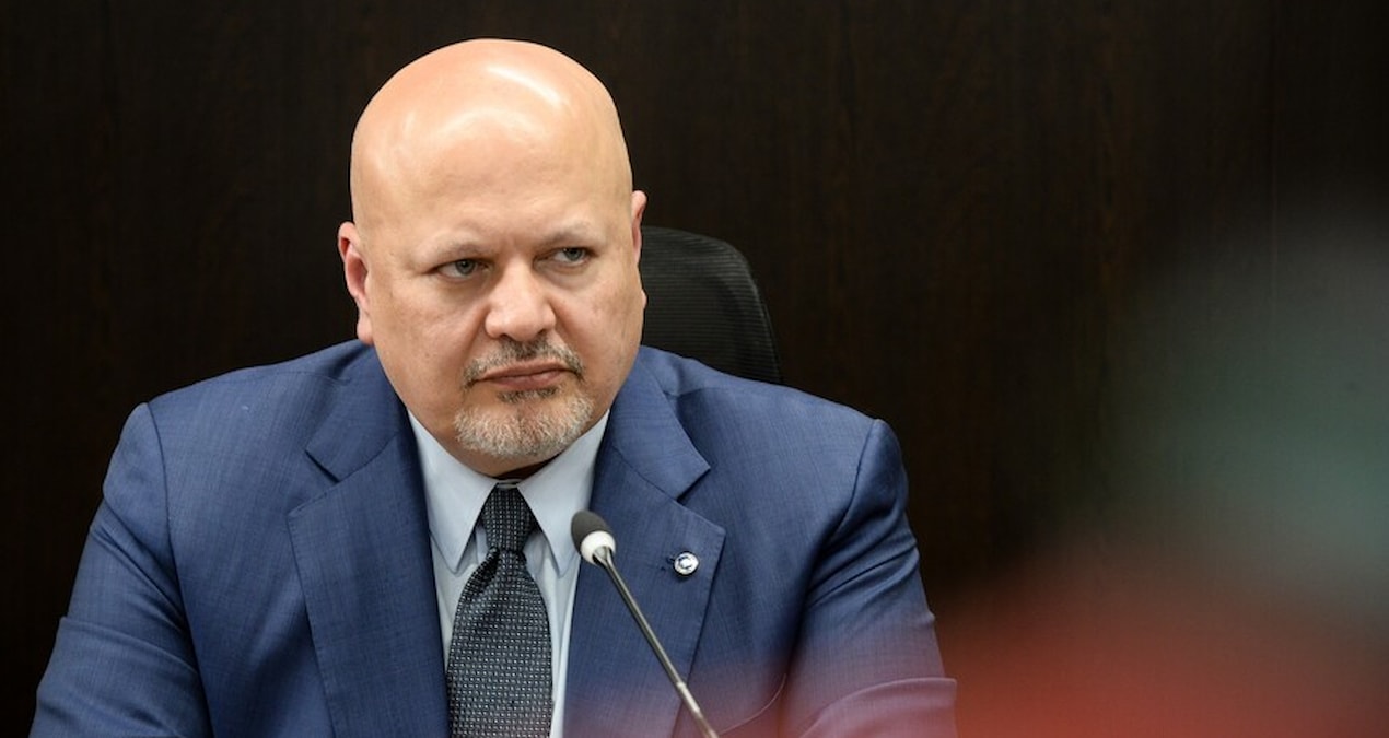 Lacivert takım elbiseli Karim Khan masada mikrofon önünde ciddi ifadeyle oturuyor