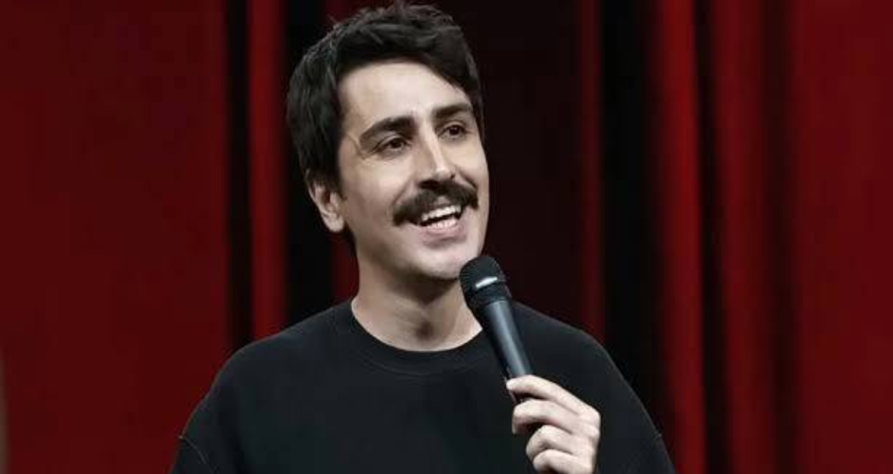 Komedyen Deniz Göktaş sahnede elinde mikrofonla gülümserken
